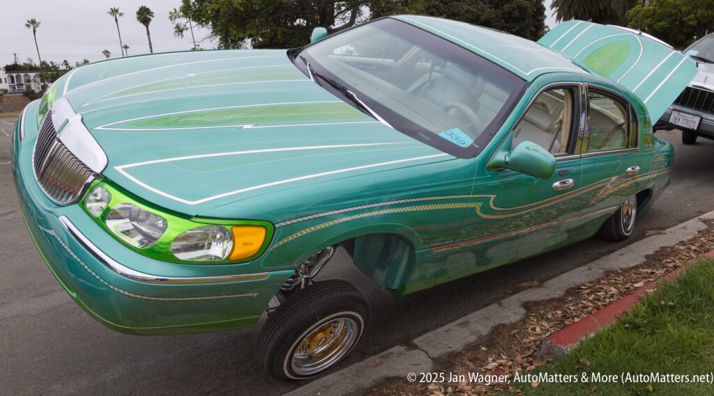 A green custom lowrider car with detailed paint, raised front suspension, and open hood is parked on the street.
