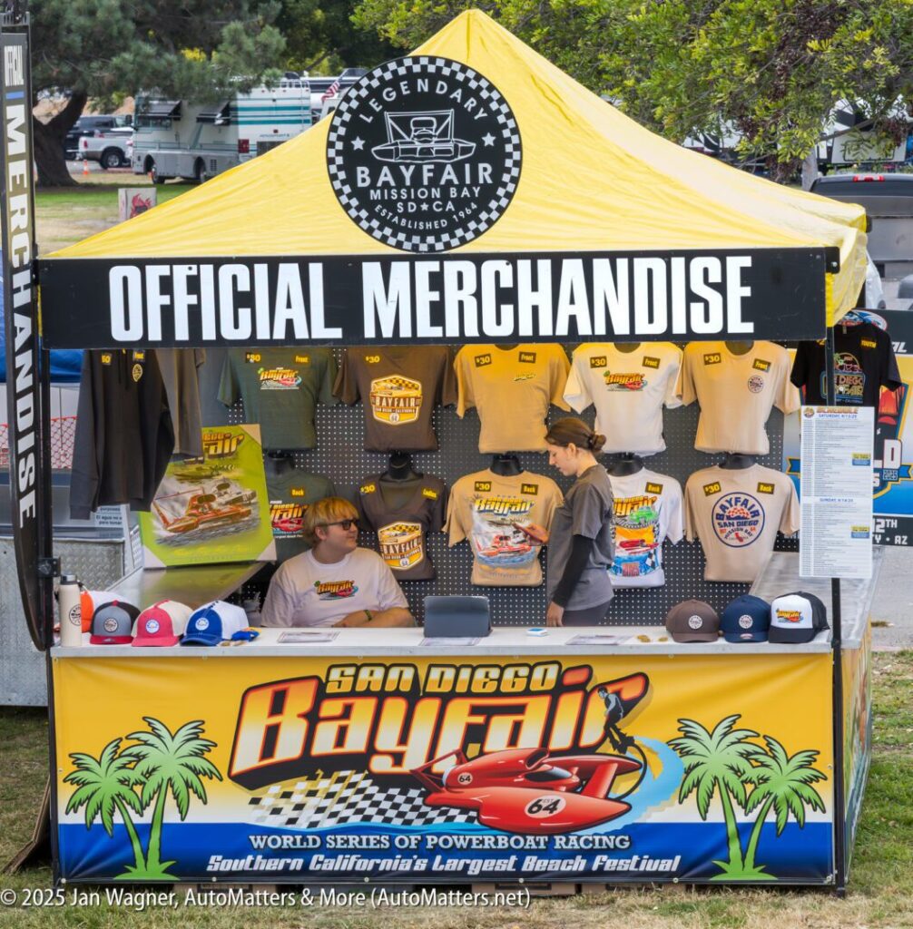 An outdoor booth with a yellow and black tent labeled "Official Merchandise" selling San Diego Bayfair event shirts, hats, and other items. Two people are working inside.