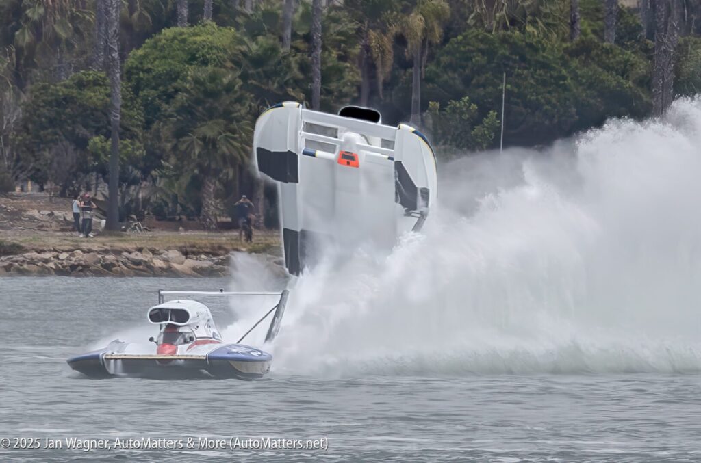 A speedboat flips over on the water, creating a large splash, while another boat passes nearby; trees and spectators are visible in the background.