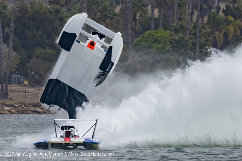 A hydroplane boat flips into the air on a lake, creating a large spray of water; another boat passes in the foreground.