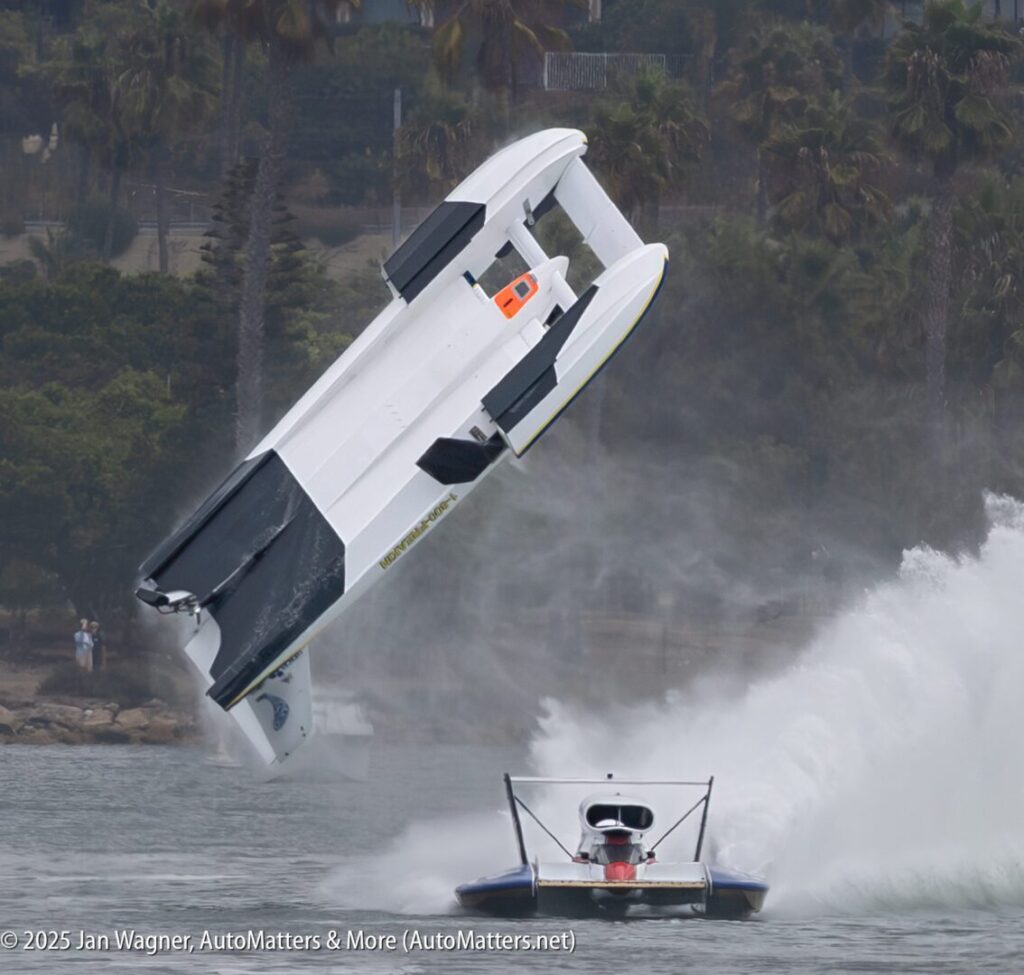 A hydroplane boat flips into the air above the water while another boat speeds past, creating a large spray; palm trees and spectators are visible in the background.