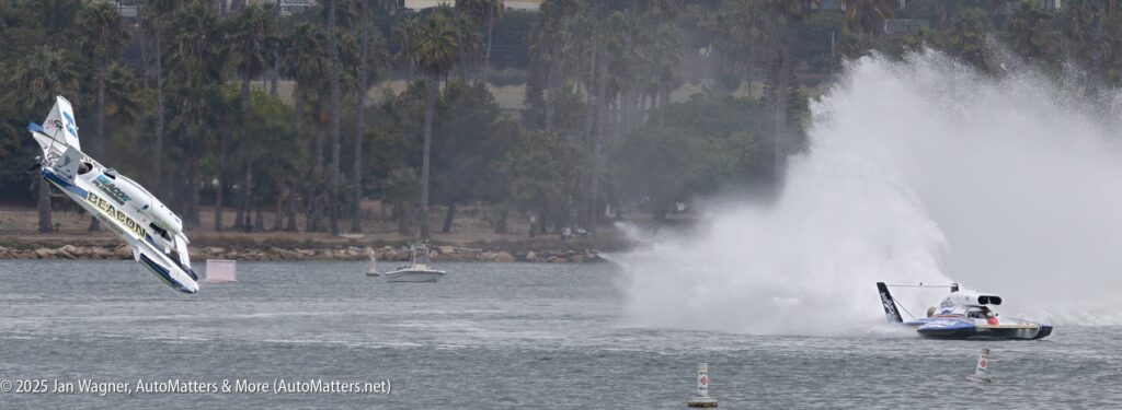 A hydroplane boat flips into the air while another boat speeds away, creating a large spray of water during a race on a river with trees in the background.