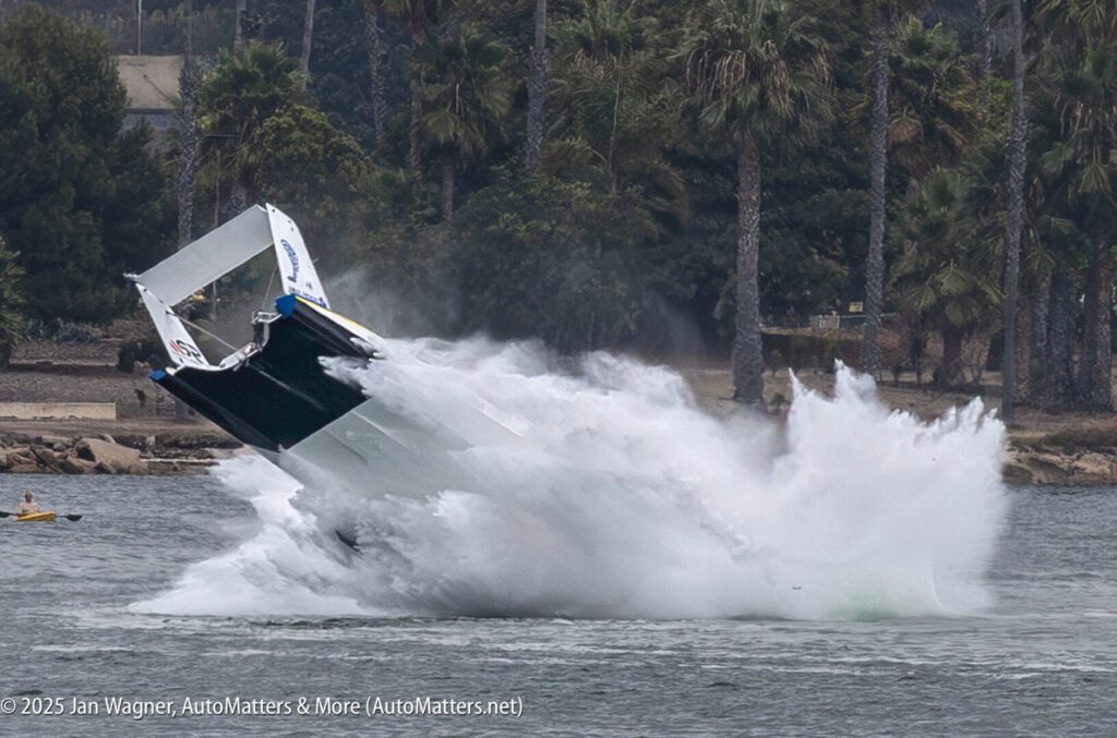 A speedboat crashes and flips upward, creating a large splash in the water with trees and shoreline in the background.