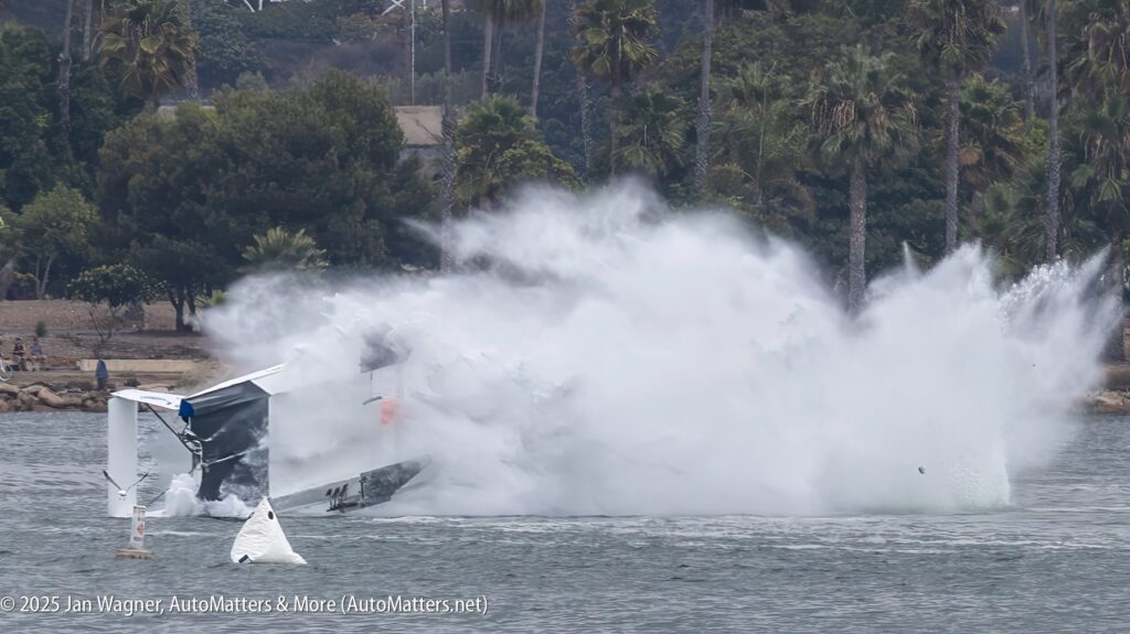 A hydroplane crashes into the water, creating a large splash and spray, with trees and people visible in the background.