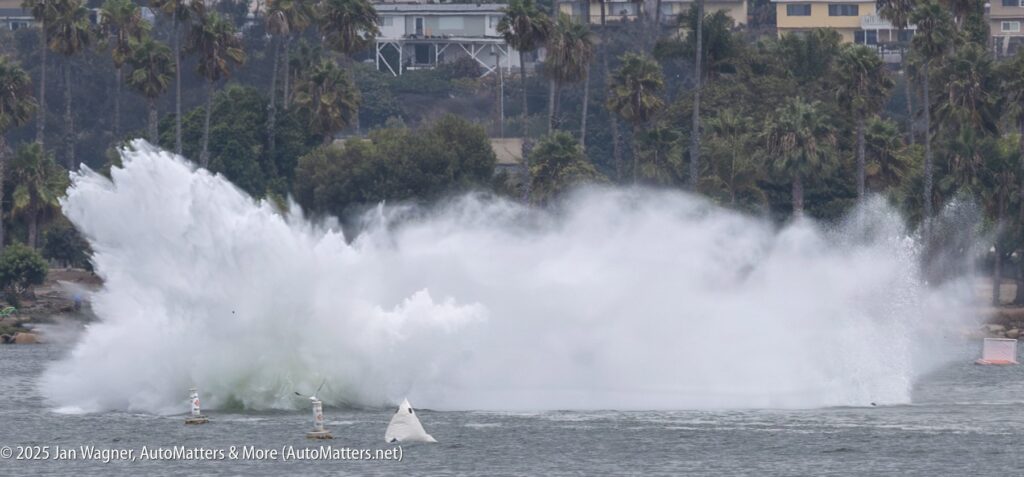 A large water splash rises from a bay, obscuring the view of any object at the center; palm trees and buildings are visible in the background.