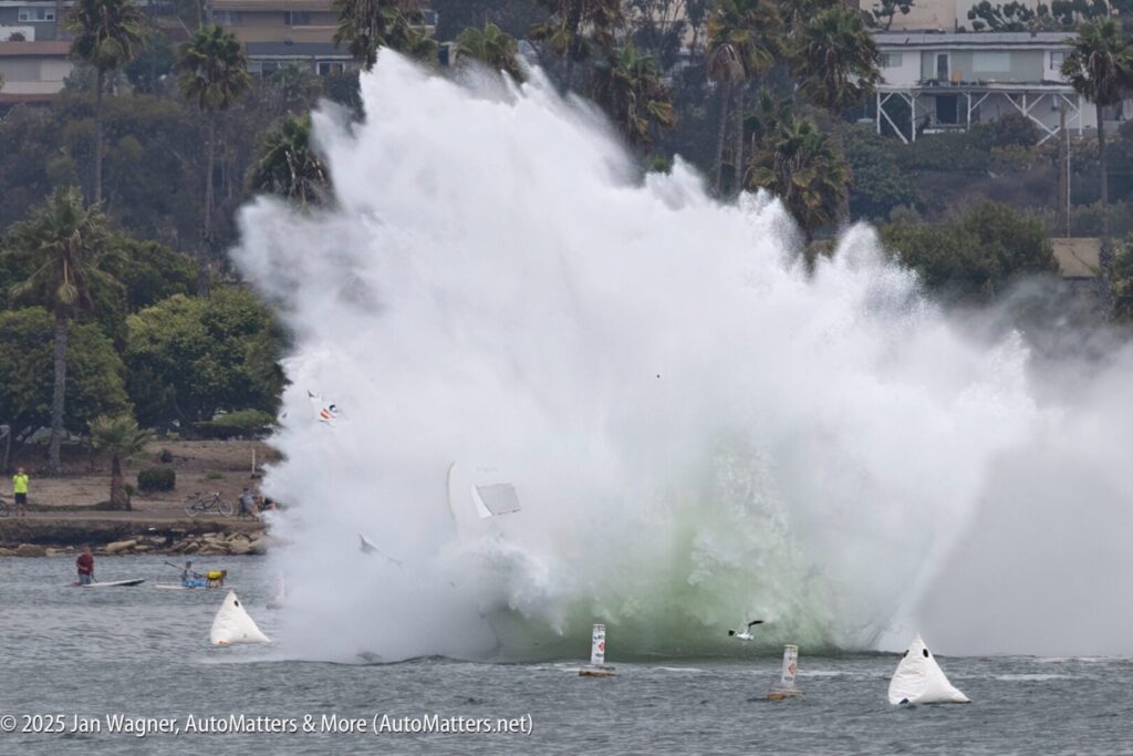 A large splash and debris are visible as a hydroplane crashes during a boat race near the shoreline, with spectators and palm trees in the background.