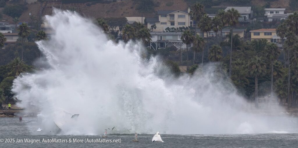 A large plume of water sprays into the air as a hydroplane crashes into a bay, with houses and palm trees visible in the background.