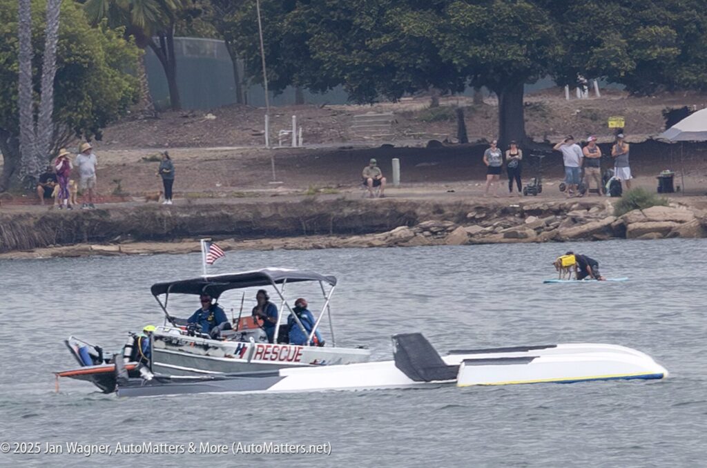 A rescue boat assists an overturned speedboat on a body of water, while people observe from the shore and one person paddles a board nearby.