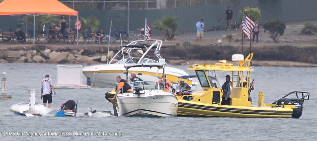 Several boats, including a yellow rescue vessel, are gathered on the water as people attend to a small capsized boat near a shoreline with spectators.