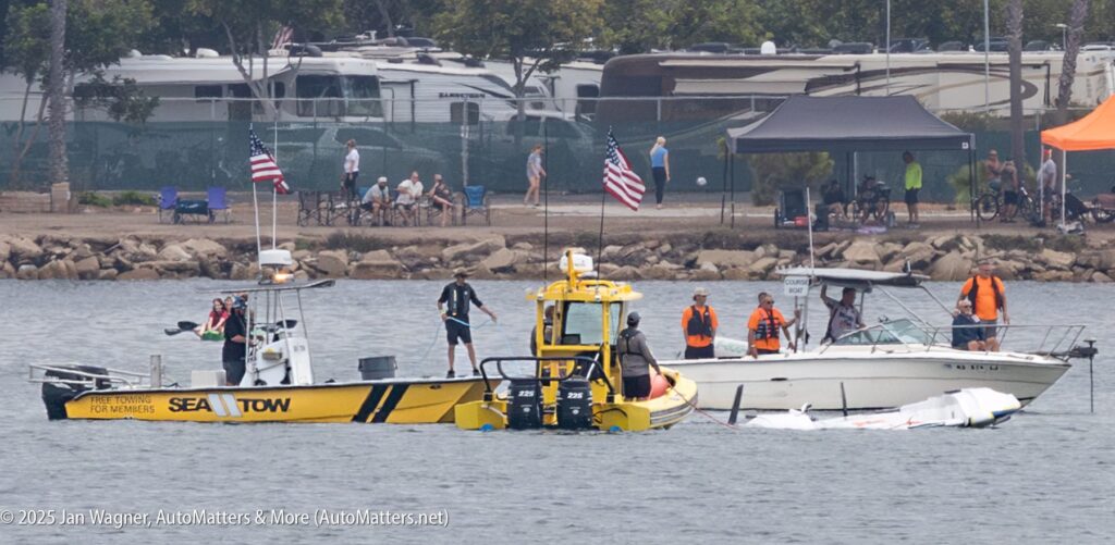 A yellow Sea Tow boat assists a white motorboat and wreckage in the water; several people in life vests are onboard, with U.S. flags displayed. RVs and onlookers are visible in the background.
