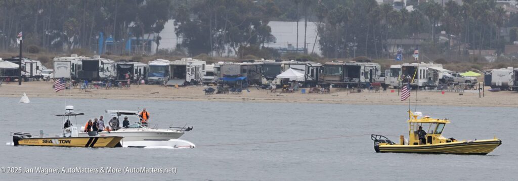 A Sea Tow boat is towing a stranded motorboat near the shore, with several people onboard; RVs and palm trees are visible in the background.