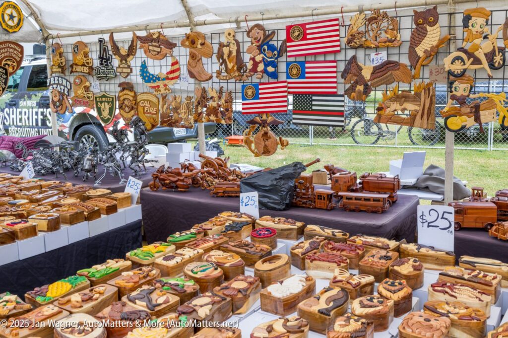 A market stall displays numerous wooden crafts, including boxes, animal figures, wall plaques, and metal sculptures, all arranged on tables under a tent.