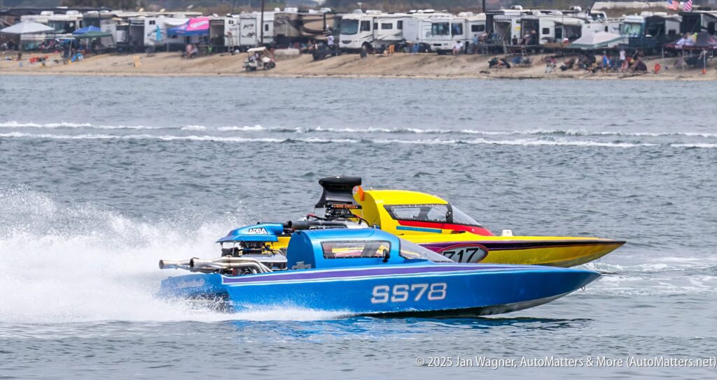 Two speedboats, one blue (S578) and one yellow (317), race side by side on a body of water, with spectators and RVs visible along the shoreline in the background.