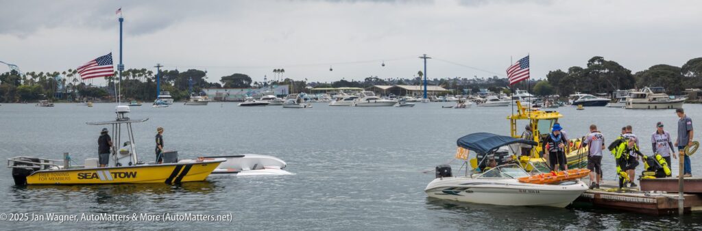 A group of people in uniforms stand on a dock next to two boats, one marked "SEA TOW for emergencies," with a marina and several boats in the background.