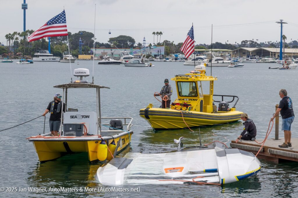 Two rescue boats assist with the recovery of a capsized sailboat near a dock, as several people manage ropes and lines.