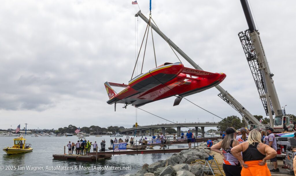 A hydroplane race boat is lifted by a crane over a dock as people watch by the water; boats and a bridge are visible in the background.