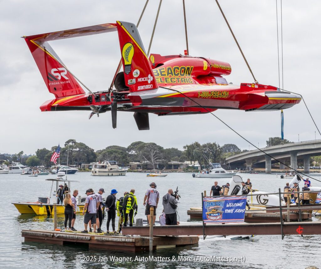 A red hydroplane race boat labeled "Beacon Electric" is being lifted by a crane over a dock, with people and boats visible in the background.