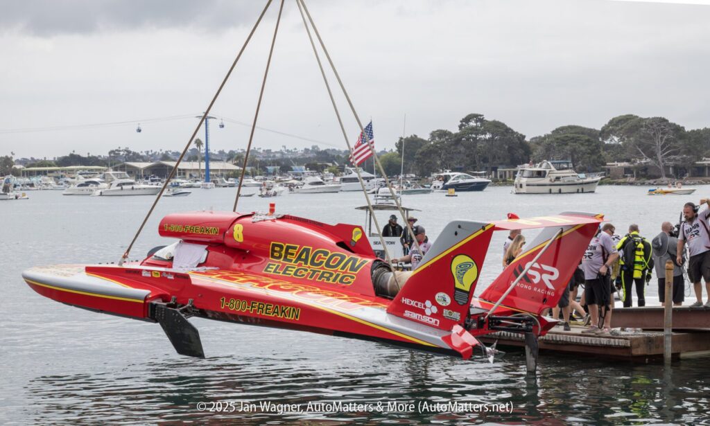 A red hydroplane boat labeled "Beacon Electric" is being lifted by a crane near a dock, with people and boats visible in the background.