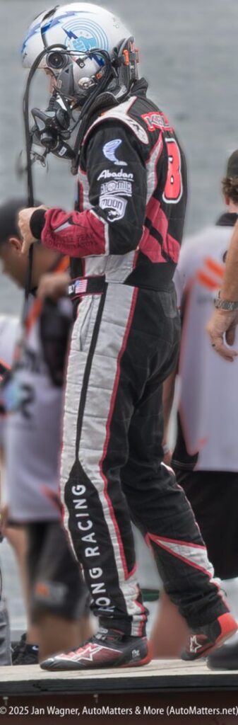 A race car driver wearing a helmet and racing suit stands next to a team, preparing for an event.