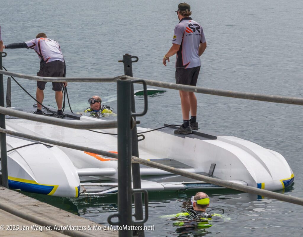 Three people in water and two standing on a dock assist with an overturned white boat at the edge of a body of water.
