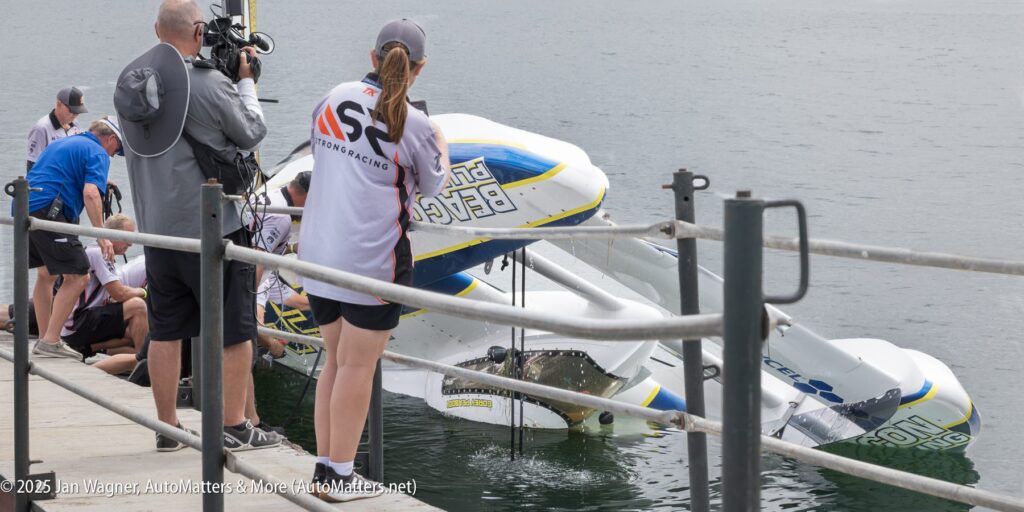 A hydroplane boat tipped on its side in the water as crew members and photographers gather on a dock nearby.