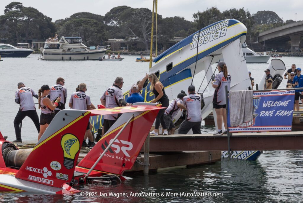 Several people work together to lift and right an overturned speedboat at a dock during a boat racing event, while others watch from nearby boats and a banner welcomes race fans.
