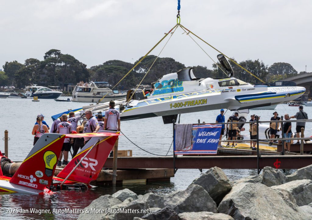 A crane lifts a hydroplane boat labeled “Go3One.com” over a dock while people in racing gear and spectators watch near the waterfront.
