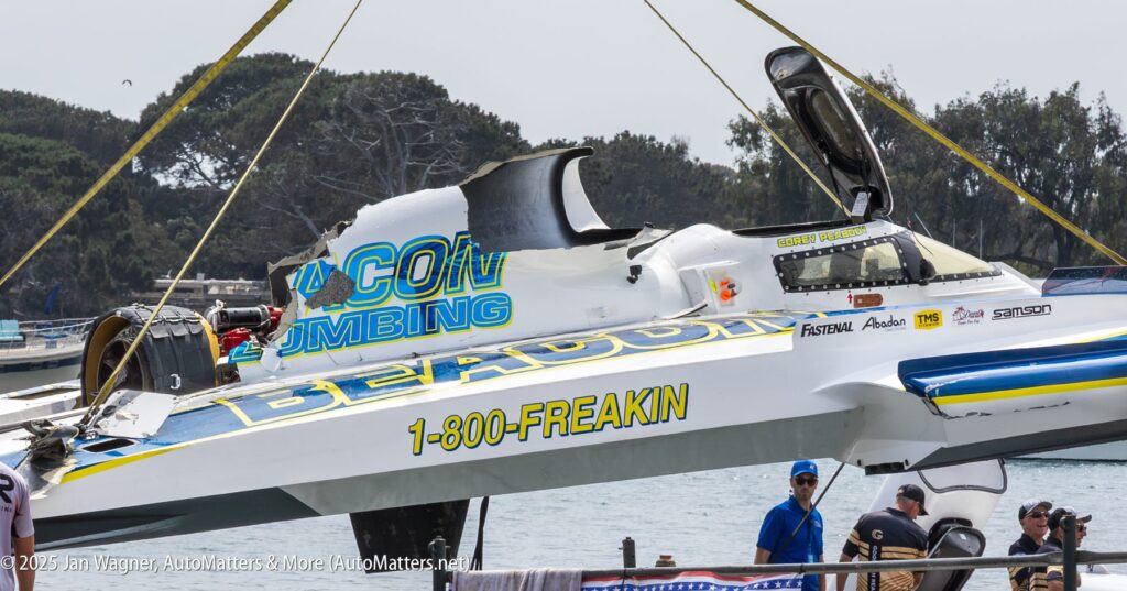 A damaged hydroplane boat labeled "1-800-FREAKIN" is lifted by a crane, with people and trees visible in the background.