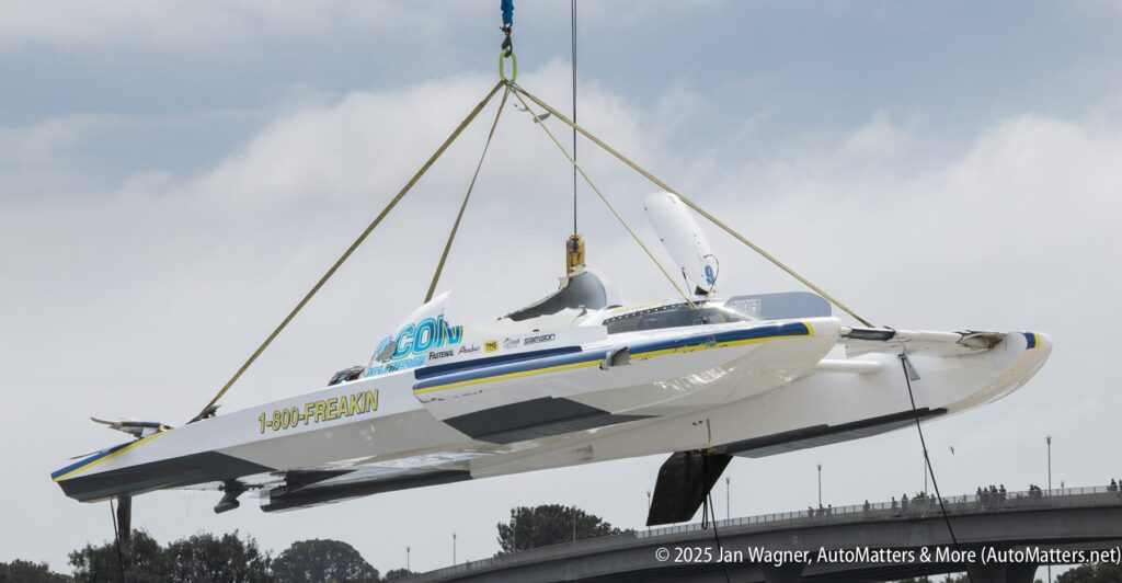 A hydroplane powerboat is suspended by a crane, with straps and cables securing it, against a cloudy sky and treetops in the background.