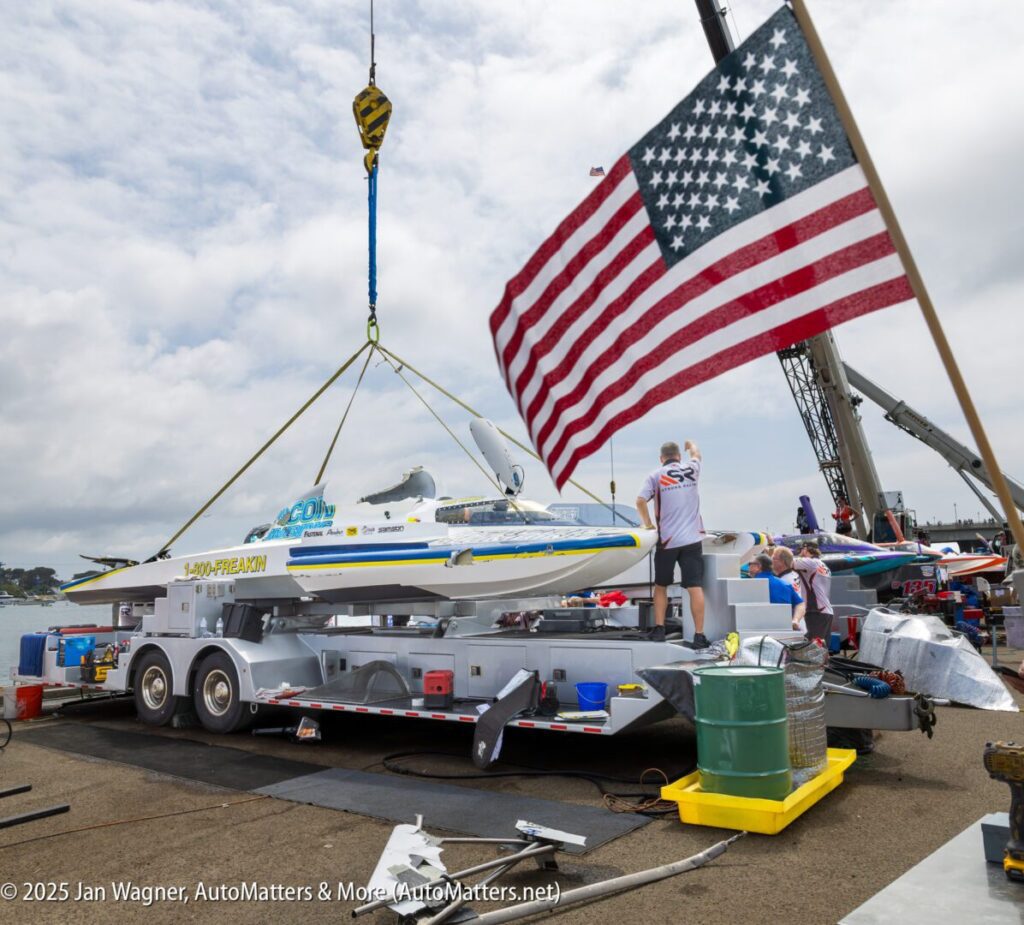 A hydroplane boat is lifted by a crane onto a trailer while people work nearby; an American flag is displayed in the foreground.