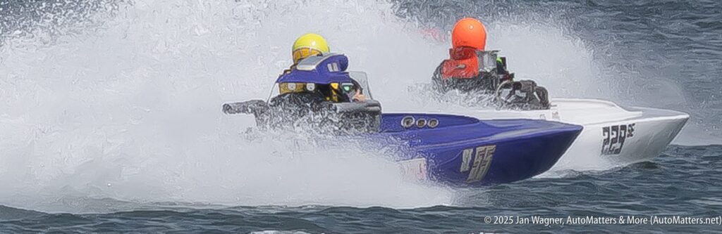 Two powerboats race side by side on water, creating large splashes; drivers wear yellow and orange helmets.