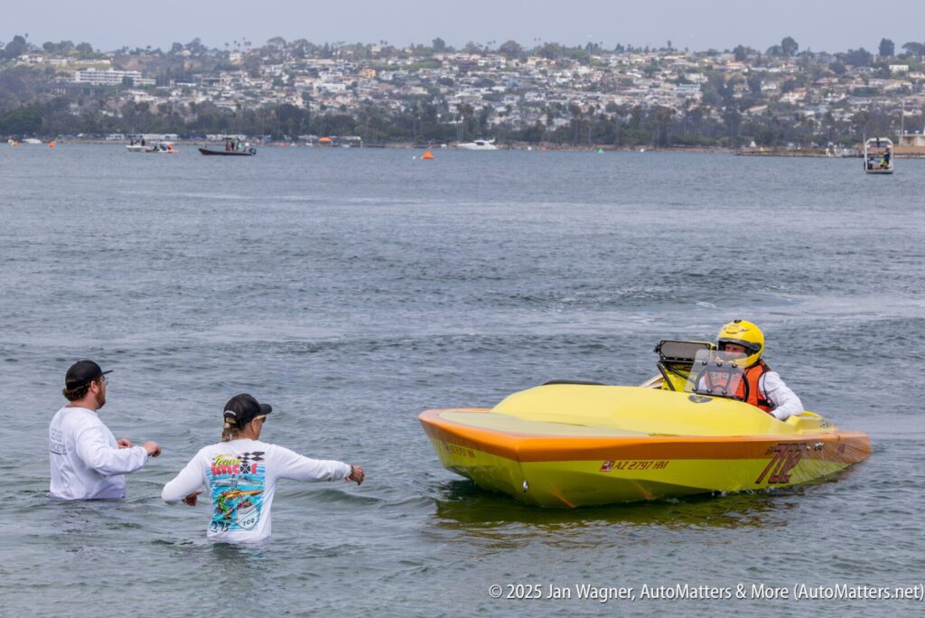 Two people in waist-deep water guide a yellow speedboat with a driver in a helmet toward the shore on a calm lake, with buildings visible in the background.