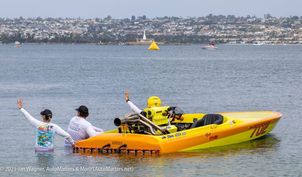 A yellow powerboat with a driver in yellow gear prepares to launch from the shore, guided by two team members standing in shallow water, with a cityscape in the background.