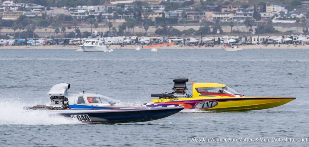 Two powerboats race side by side on the water, with a coastal town and parked vehicles visible in the background.