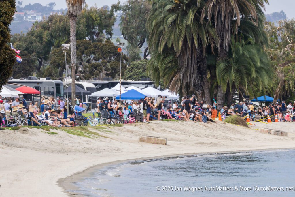 A large crowd gathers on the sandy shore near the water, with people sitting in chairs, under umbrellas, and surrounded by palm trees and RVs.
