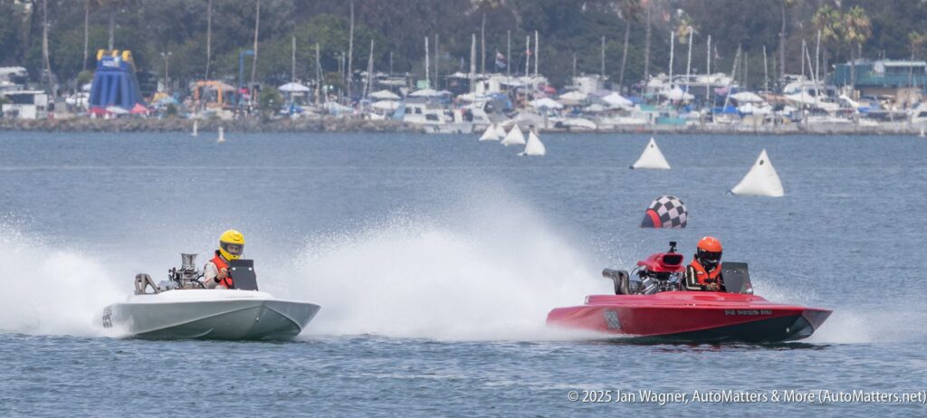 Two speedboats, one white and one red, race on the water with drivers in helmets; the red boat's driver holds a checkered flag. A marina and trees are visible in the background.