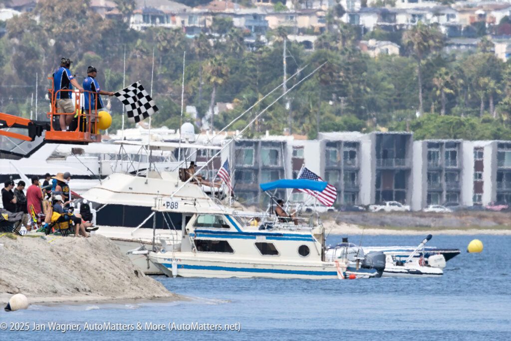 A person waves a checkered flag from a lift beside a docked boat, with people watching and buildings visible in the background.