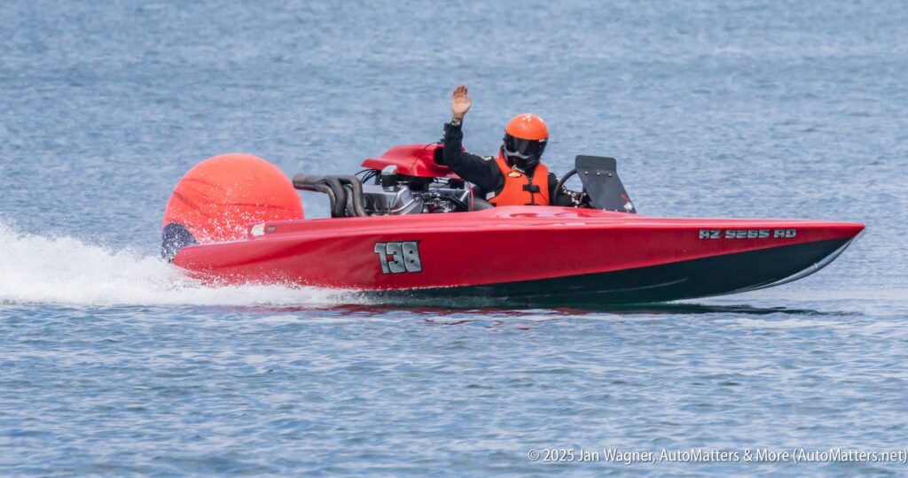 A person wearing an orange helmet and life vest waves while driving a red speedboat on the water.