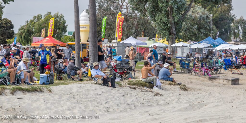 A crowd of people sit on chairs and stand on a sandy area near vendors and tents at an outdoor event.