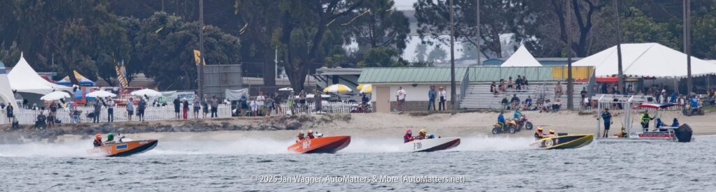 Five speedboats race on water near a sandy shore with spectators, tents, and trees in the background under an overcast sky.