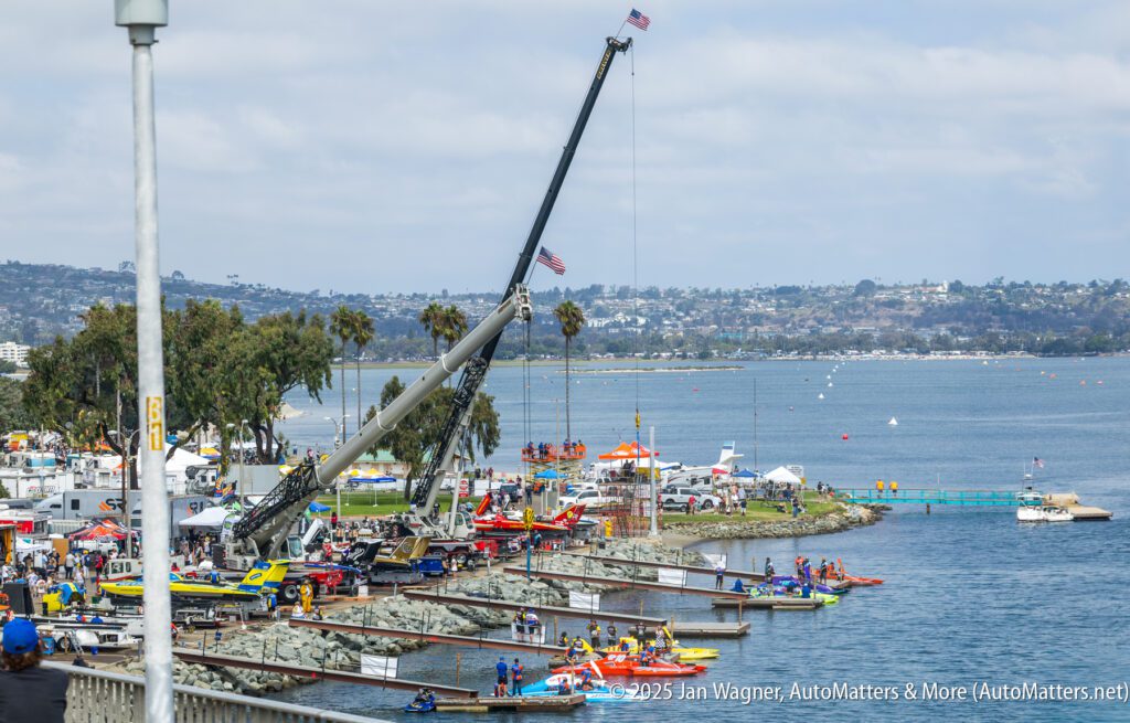 A crane lifts an object near colorful boats docked at a lakeside event, with tents, palm trees, and people along the shoreline. A cityscape and hills are visible in the background.