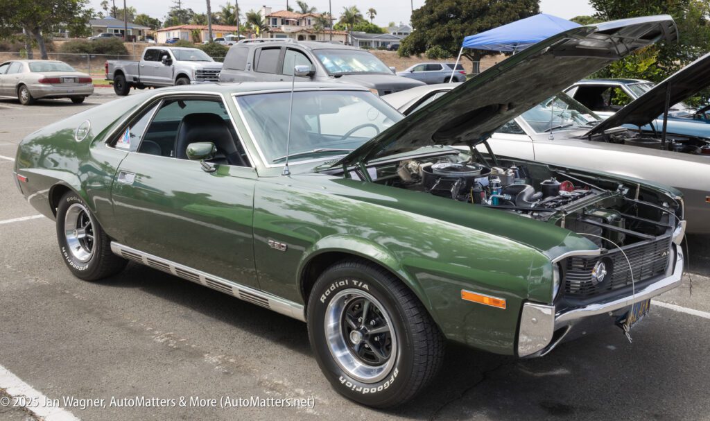 A green classic AMC AMX car with its hood open, parked at a car show in an outdoor lot with other vehicles in the background.