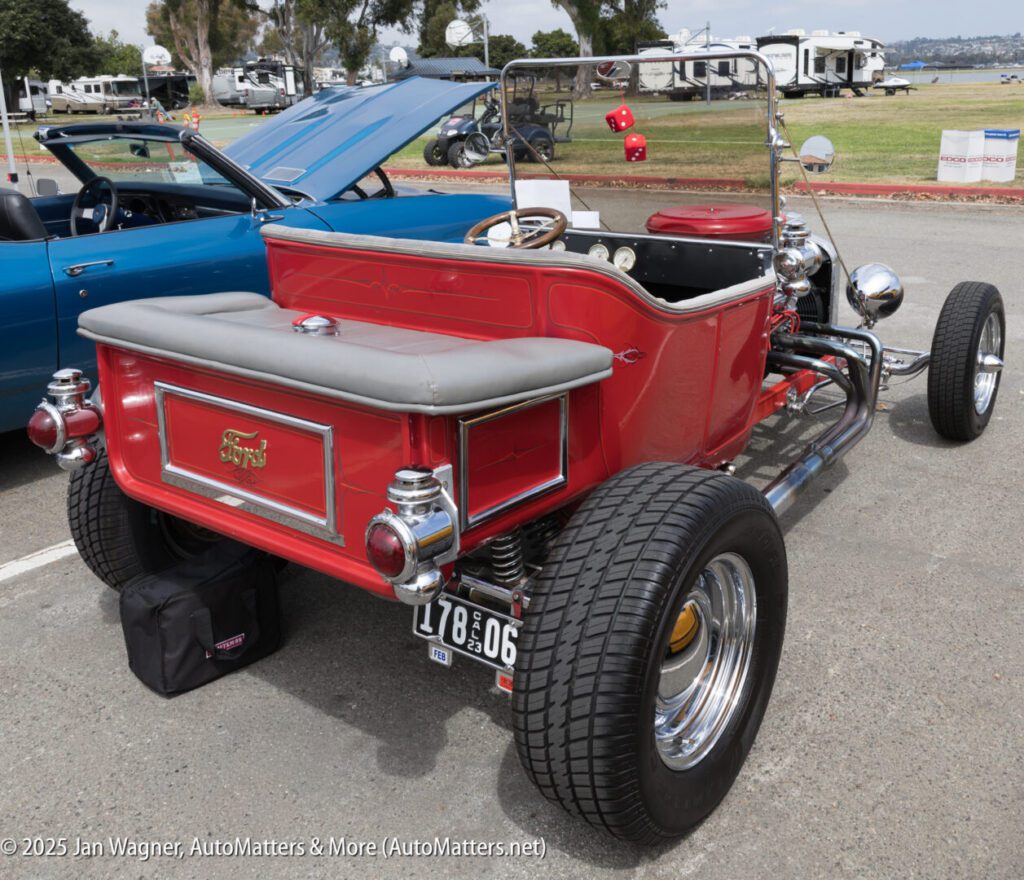 A classic red Ford T-bucket hot rod with chrome accents is parked at a car show, viewed from the rear left side.