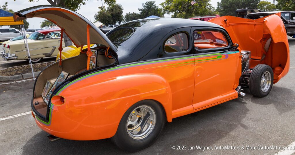 Bright orange and black classic hot rod car with the hood and trunk open, parked at an outdoor car show with other vintage cars visible in the background.