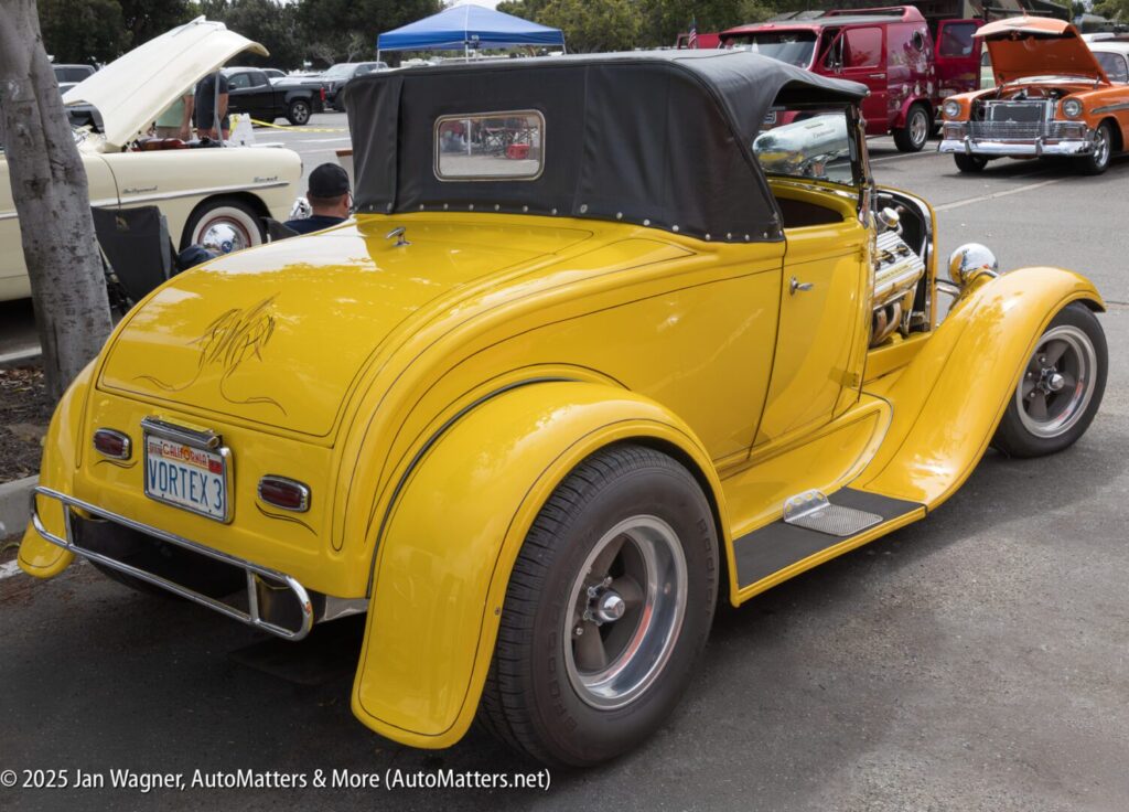 A yellow vintage convertible hot rod with a black soft top is parked at a car show, surrounded by other classic cars.