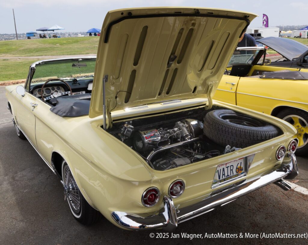 A yellow vintage convertible car with its rear hood open, revealing an engine and spare tire. The car has a "VAIR 64" license plate.