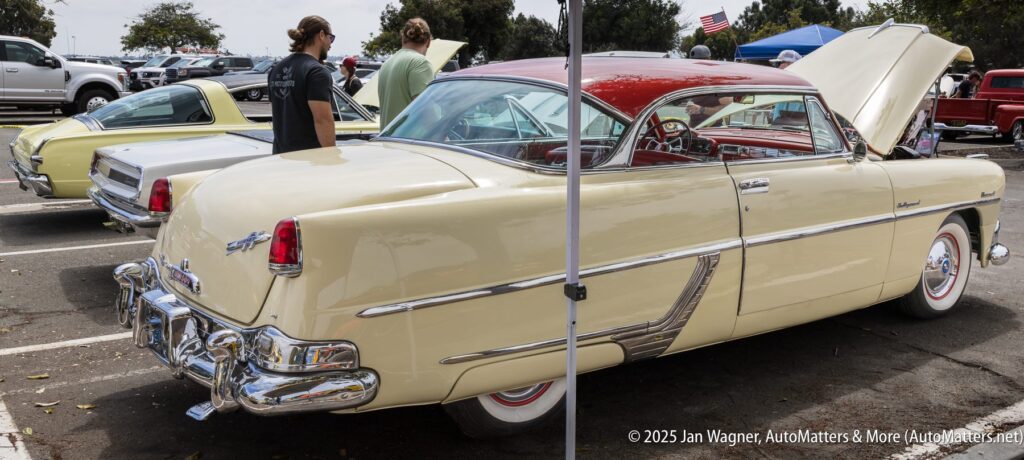 Two vintage cars are parked at an outdoor car show; one is a cream and red classic with its hood open, and two people are standing nearby examining the vehicles.