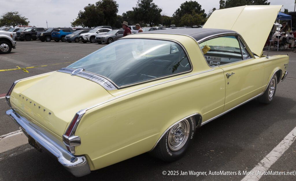 A pale yellow classic Plymouth Barracuda car with its hood open, parked at an outdoor car show.