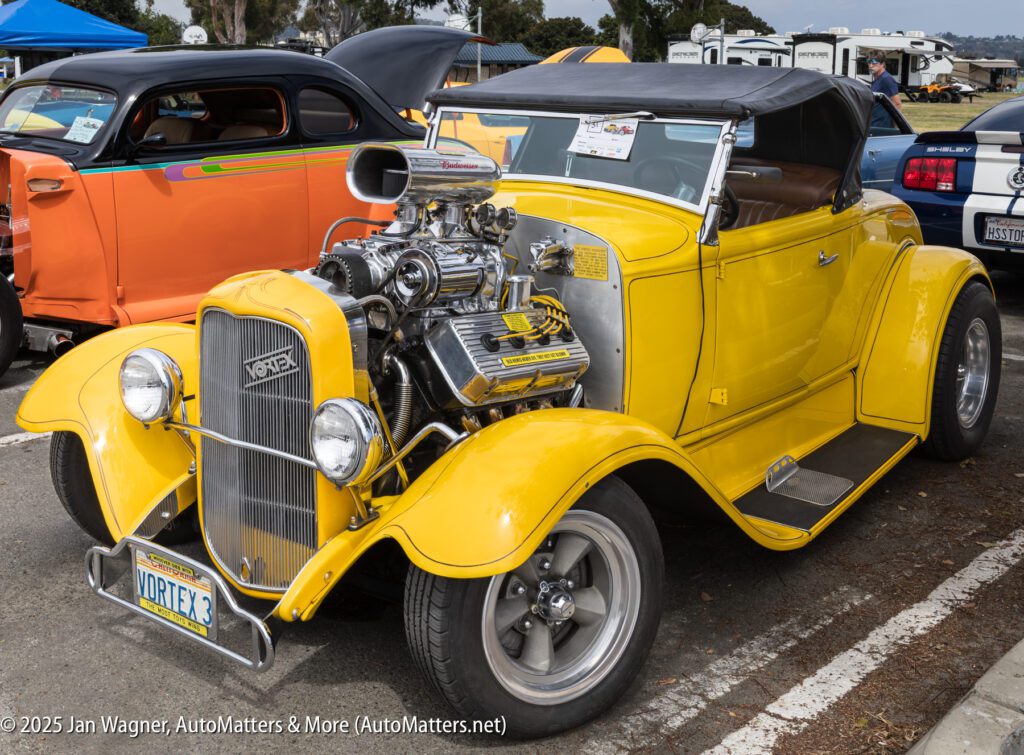 A bright yellow vintage hot rod car with exposed engine and VORTEX plate is parked at a car show; other classic cars are visible in the background.