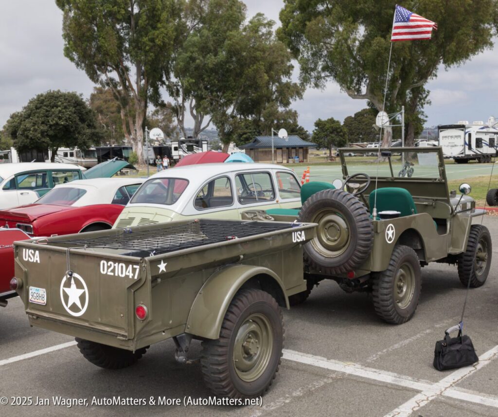 A vintage military Jeep and matching trailer with US Army markings are parked at a car show next to classic vehicles, with an American flag attached to the Jeep.